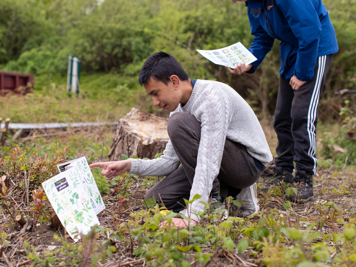 A group stood outside, a teacher giving an explanation to 3 students, one with a clipboard