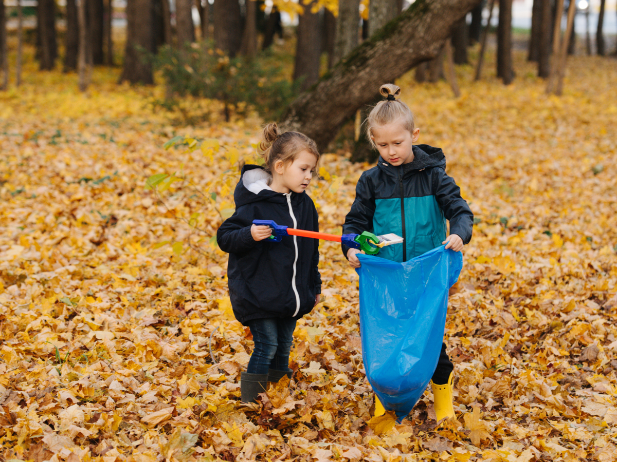 Two young children are stood in a pile of fallen leaves. One is holding a litter picker, putting some litter into the blue plastic bag that the other child is holding.
