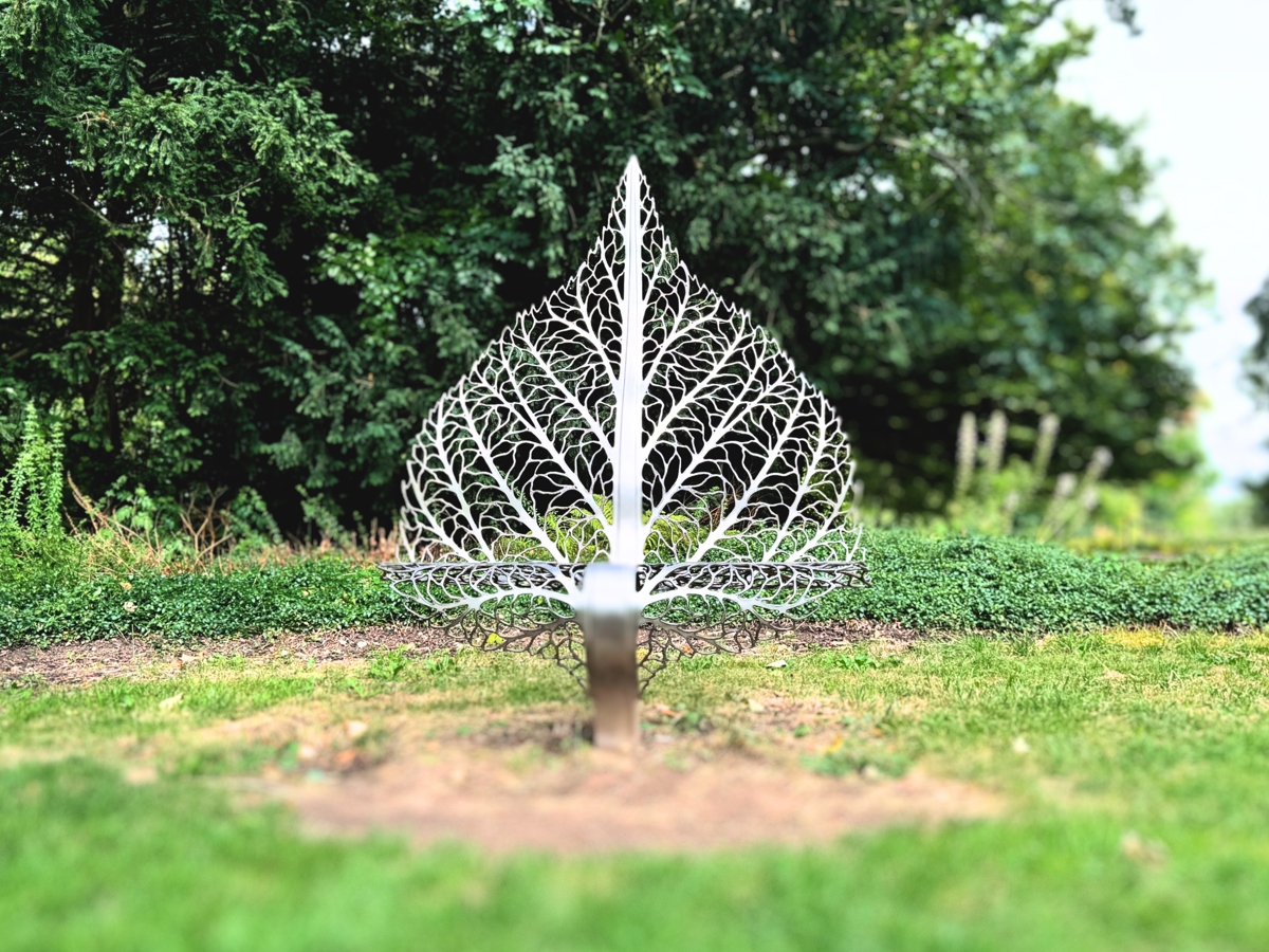 This image shows a silver metal sculpture in the shape of a large, intricate leaf. The leaf is standing upright on a patch of grass, with lush green bushes and trees in the background.
