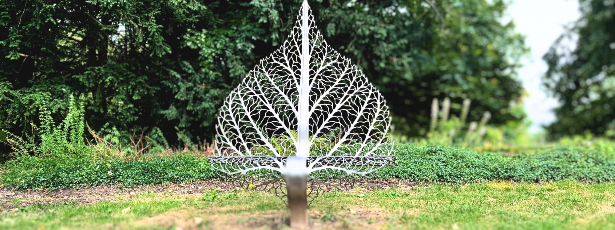 This image shows a silver metal sculpture in the shape of a large, intricate leaf. The leaf is standing upright on a patch of grass, with lush green bushes and trees in the background.