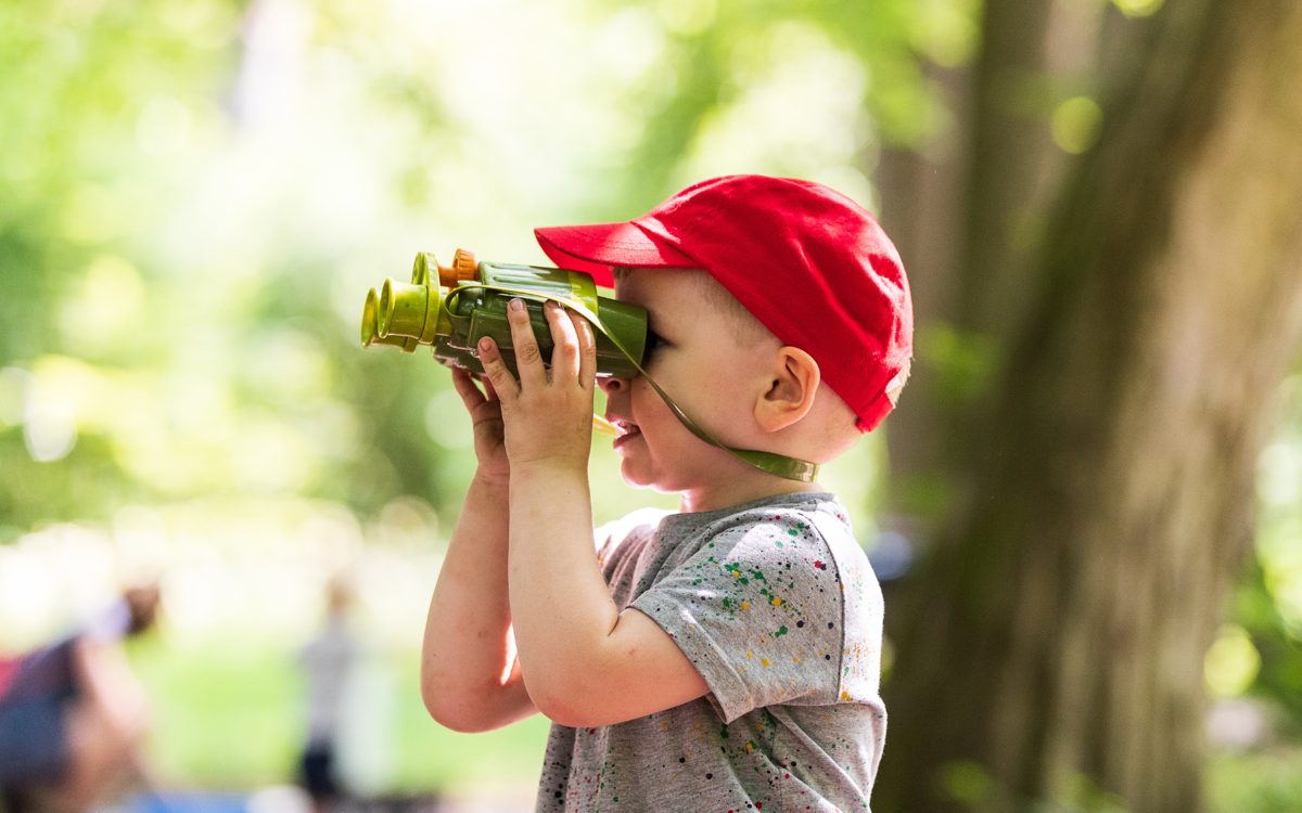 A young boy wearing a red summer hat using some binoculars in a sunny woodland