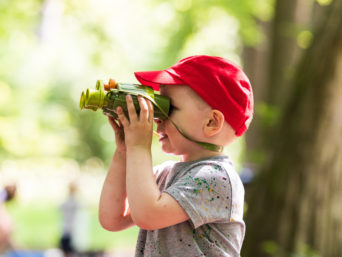 A young boy wearing a red summer hat using some binoculars in a sunny woodland