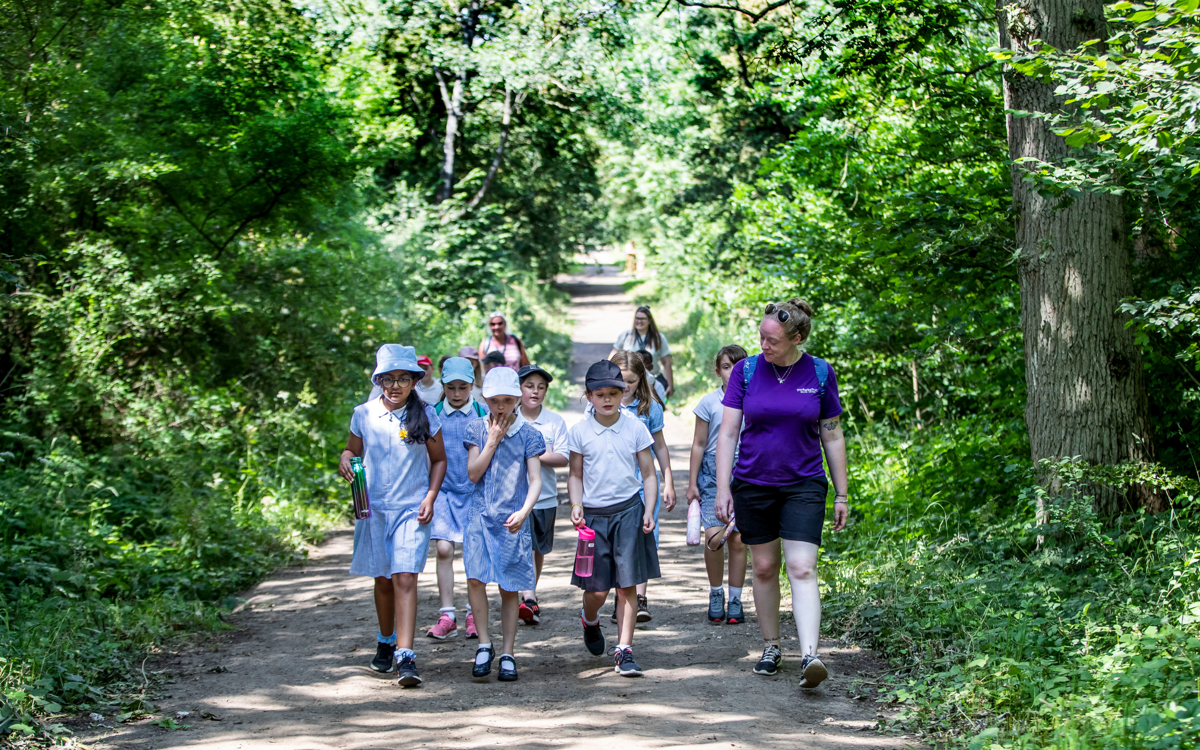 Children and a leader walk through a shady woodland on a summer's day. They are walking up a dry muddy path toward the camera. Everyone is engaging in conversation and smiling.