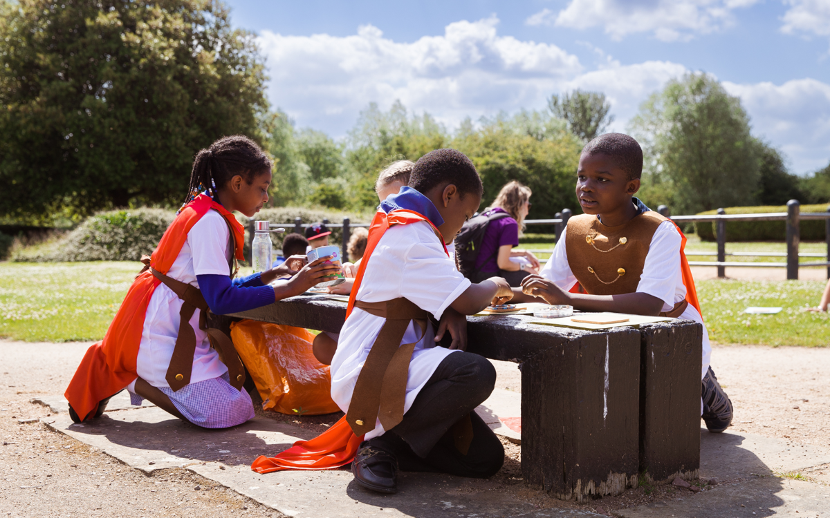 Children wearing Roman costumes engage in an activity at a bench in the sunshine