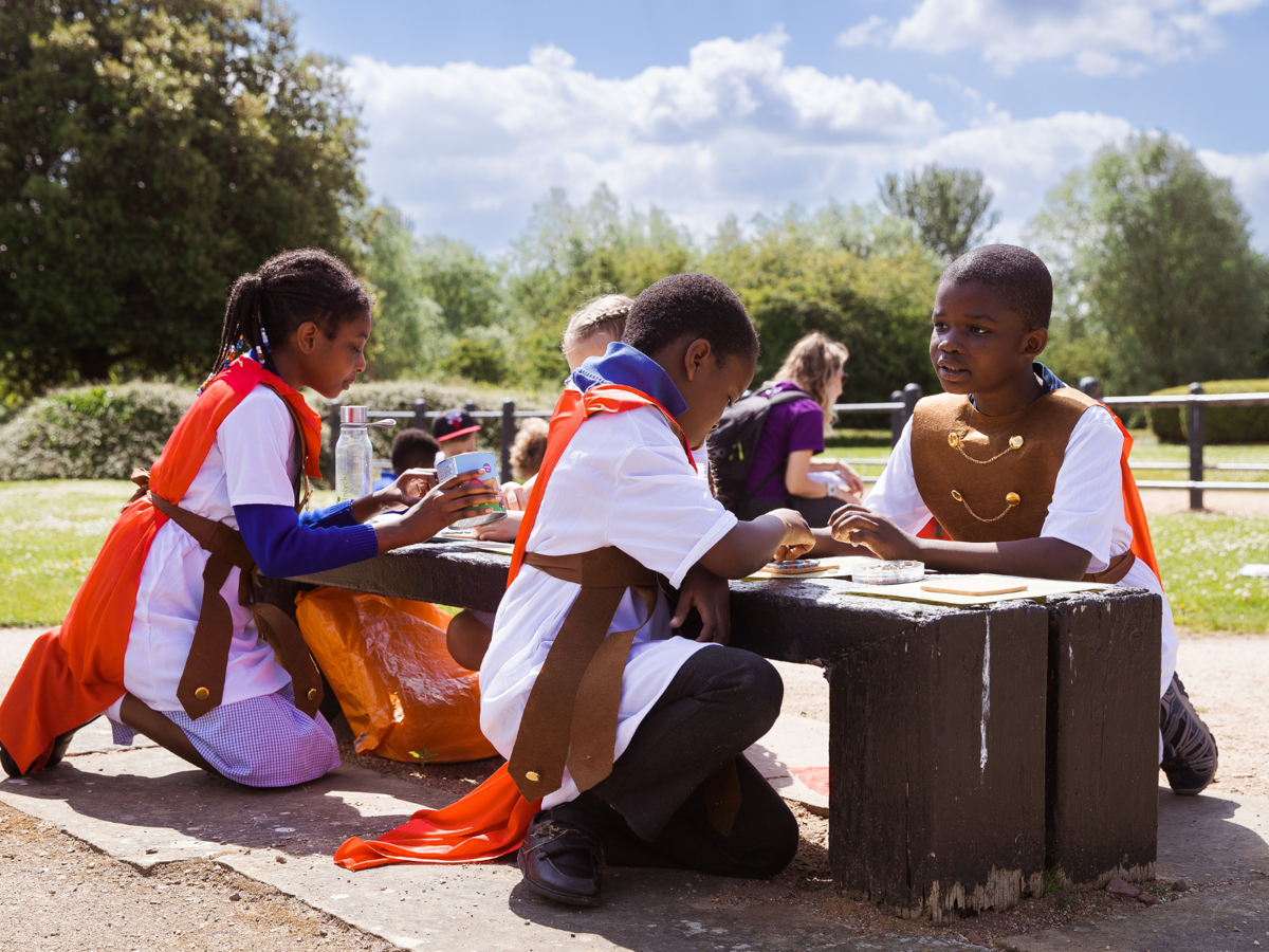 Children wearing Roman costumes engage in an activity at a bench in the sunshine