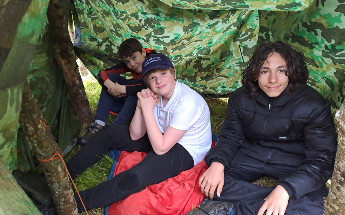 Three young boys sit under a shelter they have built using tree trunks, orange ropes and a camouflage-printed green plastic sheet