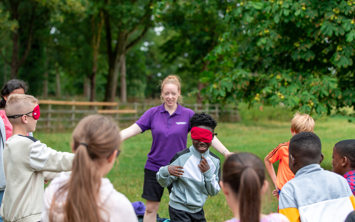 Children and a leader playing a game in a grassy parkland. Two of the children are wearing red blindfolds, running around inside a circle created by their class mates holding their arms out to their sides. 
