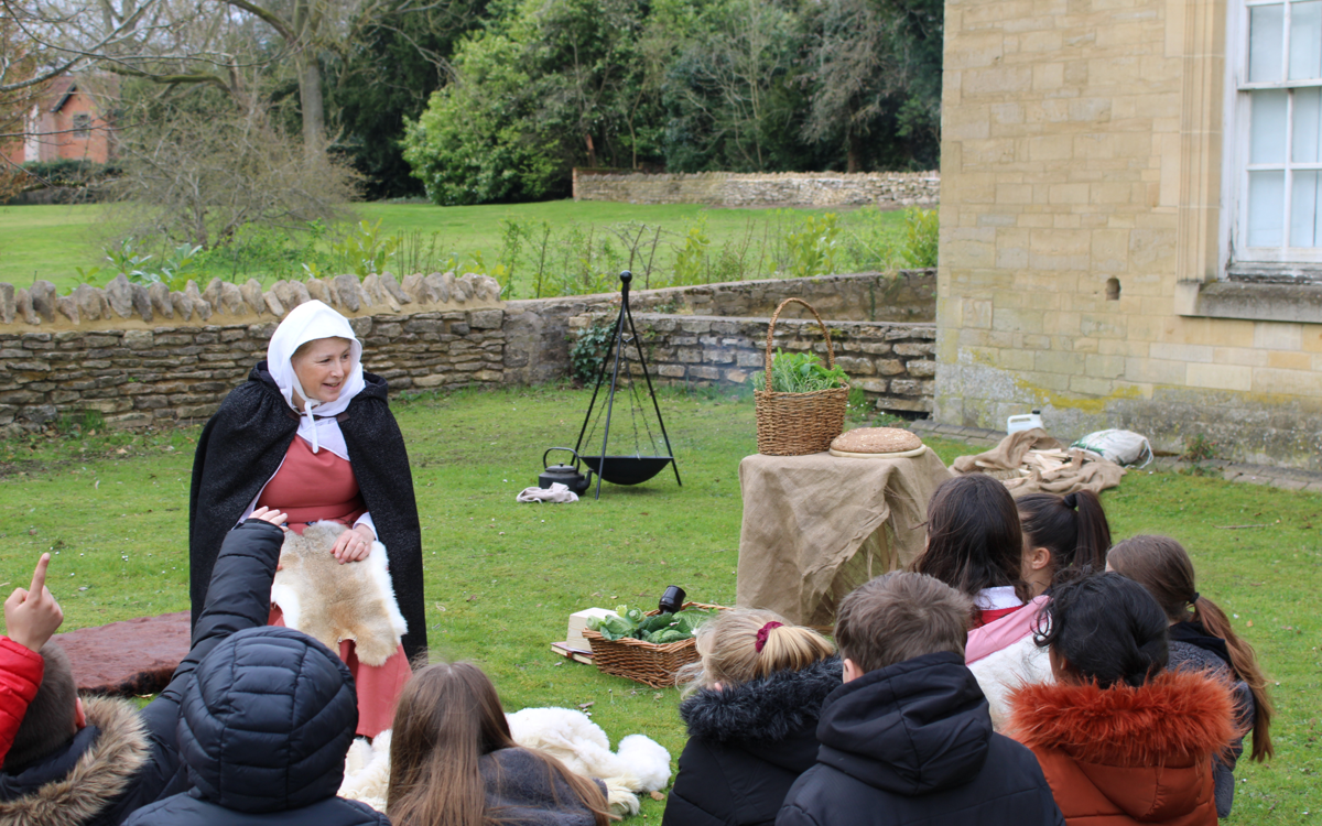 A lady in medieval costume is talking to a group of school students. They are sat on the grass out the front of an old building. A firepit and baskets of food are seen in the background.