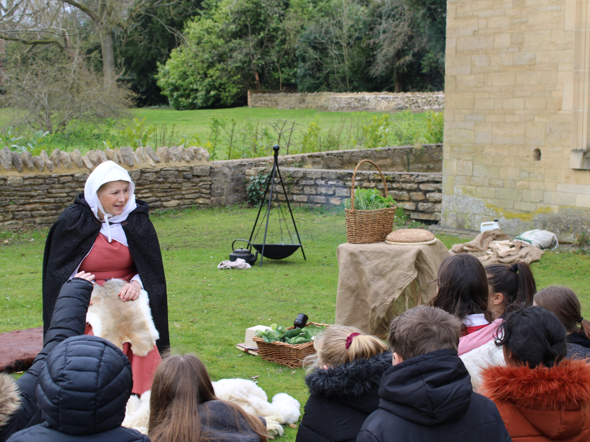 A lady in medieval costume is talking to a group of school students. They are sat on the grass out the front of an old building. A firepit and baskets of food are seen in the background.