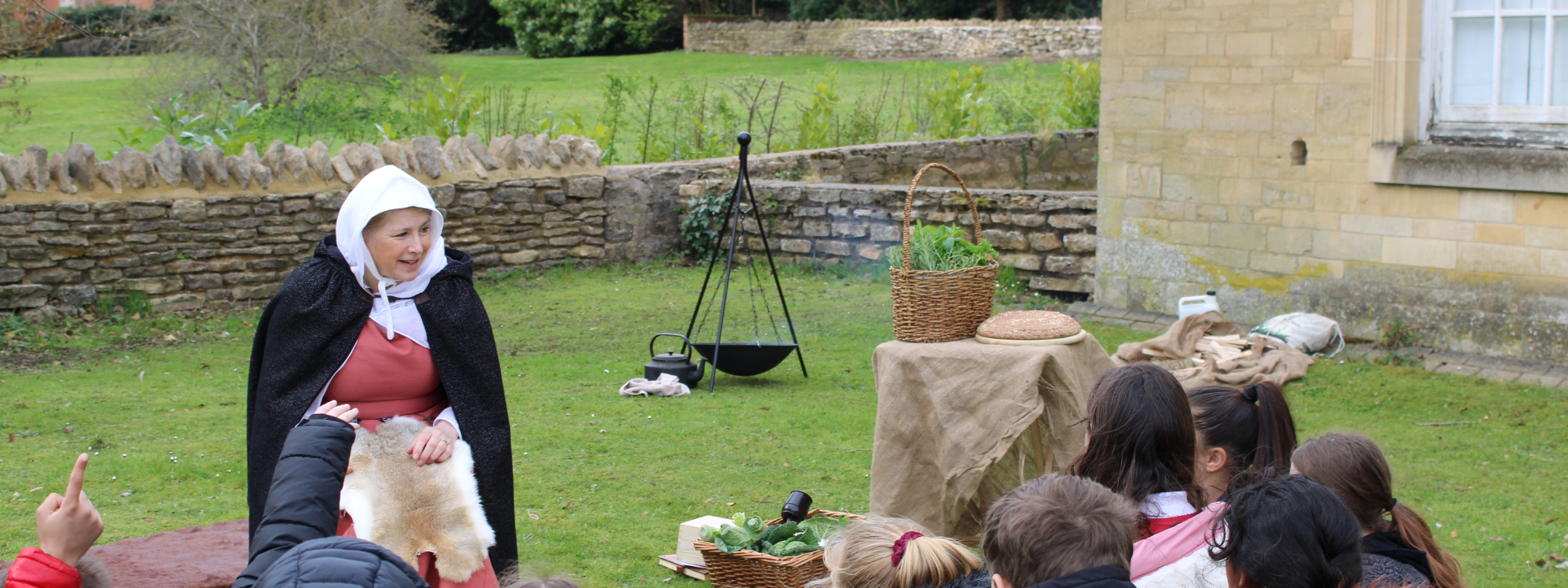A lady in medieval costume is talking to a group of school students. They are sat on the grass out the front of an old building. A firepit and baskets of food are seen in the background.