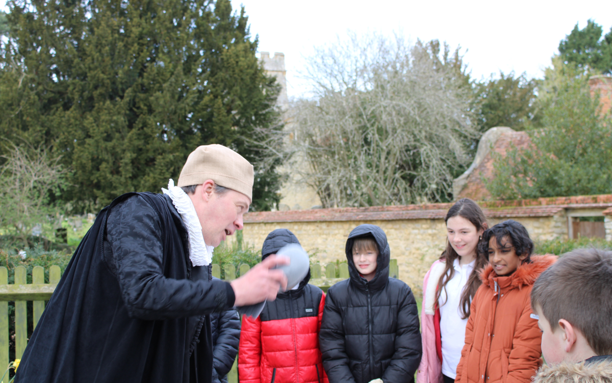 A man in medieval costume speaking to a group of school children in an outdoor medicinal garden.