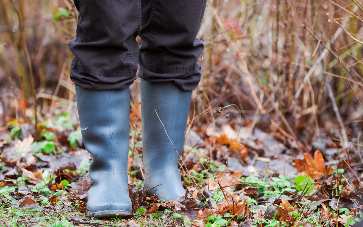A pair of legs wearing black wellies and black trousers. The person is standing on a muddy, grassy path with lots of orange and brown fallen leaves.