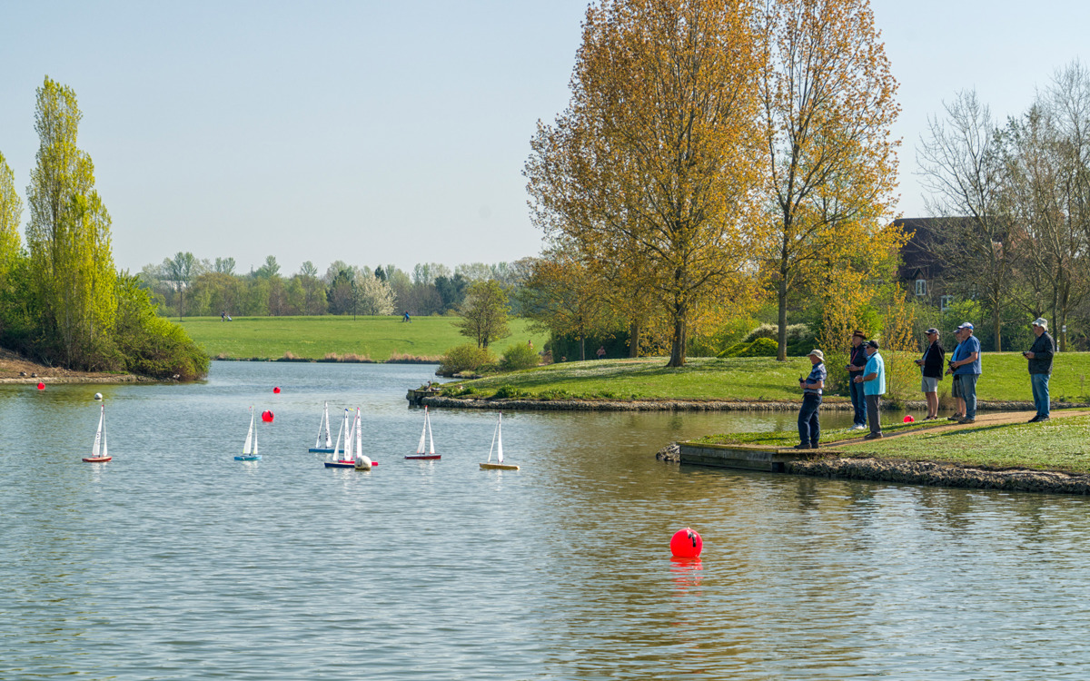 A group of people standing on the lakeside and model sailing boats in the water.