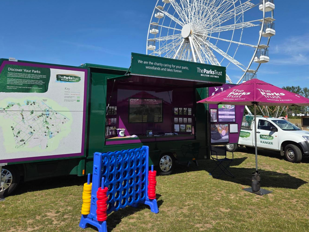 Large green community engagement van, with leaflets and posters on display, outside on a sunny day. A parasol is up, there is a giant connect 4 game out and also a Parks Trust ranger truck alongside.