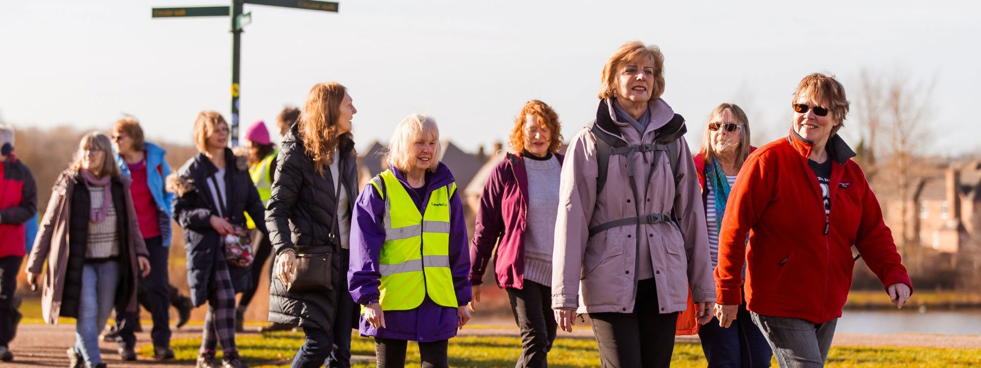A group of women walking in a parkland scene with a sign post and a lake in the background.
