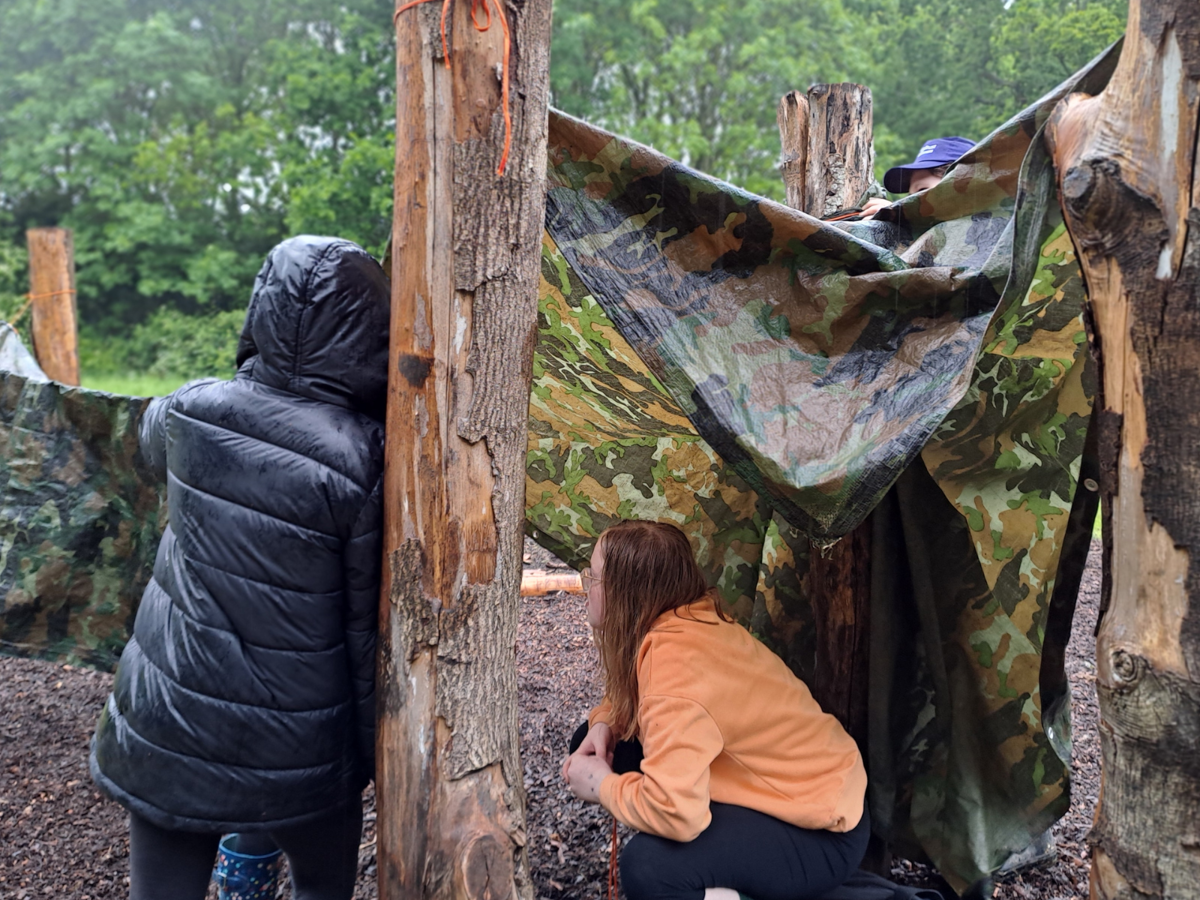 Children in the rain building shelters between tree trunks with camouflage tarpaulin