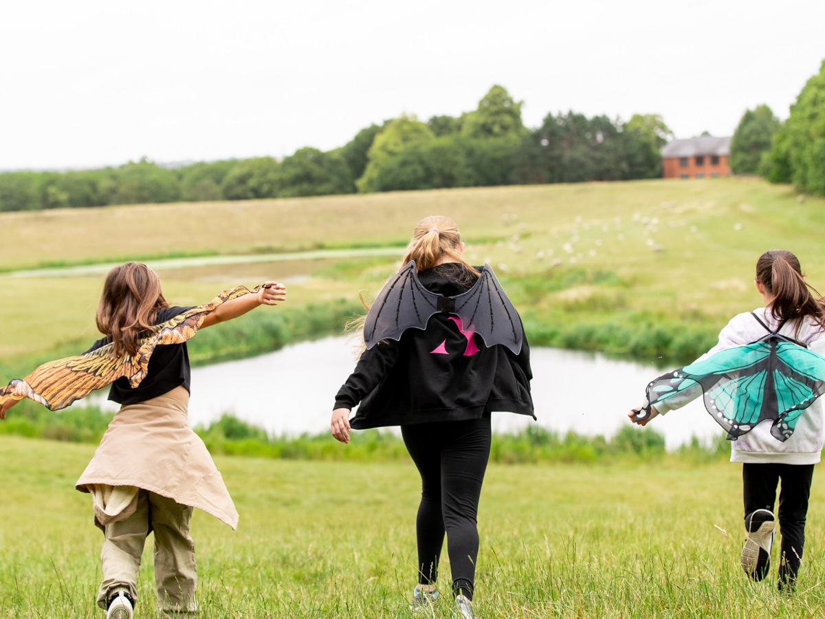 An image from behind of three girls running down a grassy hillside with a lake in the background. The girl on the left is wearing orange fabric bird wings, the middle girl is wearing fabric bat wings, and the girl on the right is wearing fabric green butterfly wings. 