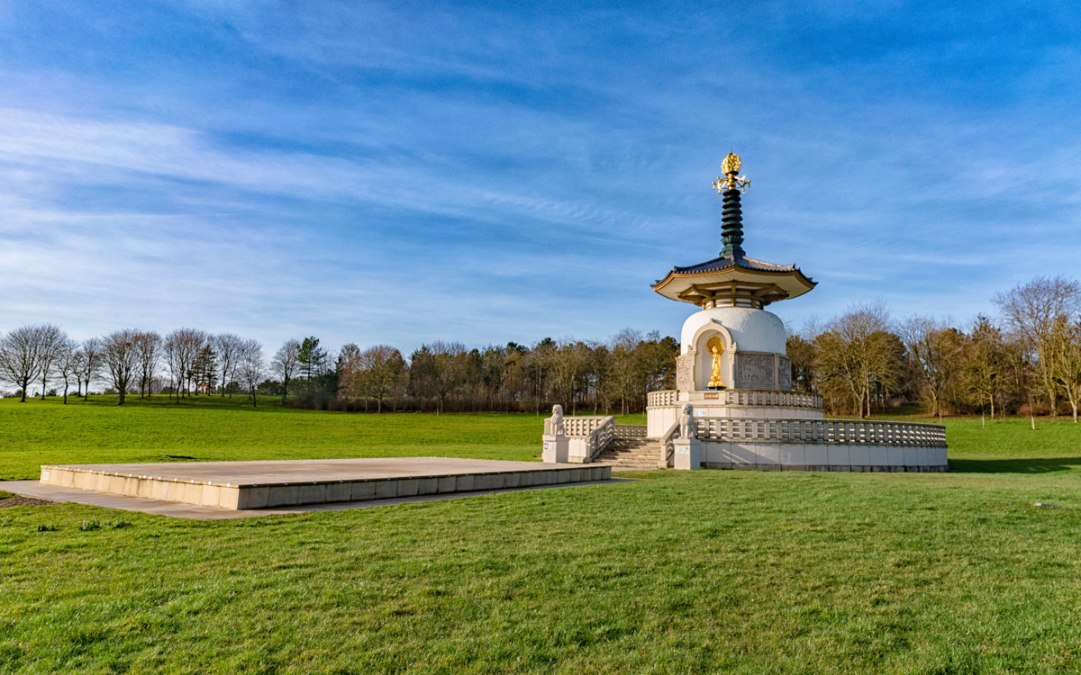 The image shows a white Buddhist stupa with a golden statue of Buddha placed in an alcove at the front. The stupa is situated in a grassy area with trees in the background under a blue sky with some clouds. The stupa features a traditional pagoda-style roof with a golden ornament at the top, and it is elevated on a platform with steps leading up to it. The overall design reflects elements of Japanese or East Asian architecture.