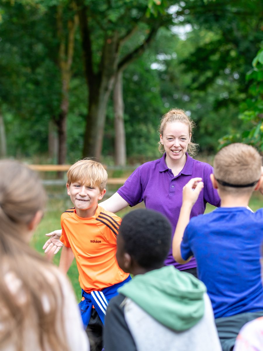 Children and a leader playing a game in a grassy parkland. Two of the children are wearing red blindfolds, running around inside a circle created by their class mates holding their arms out to their sides. 