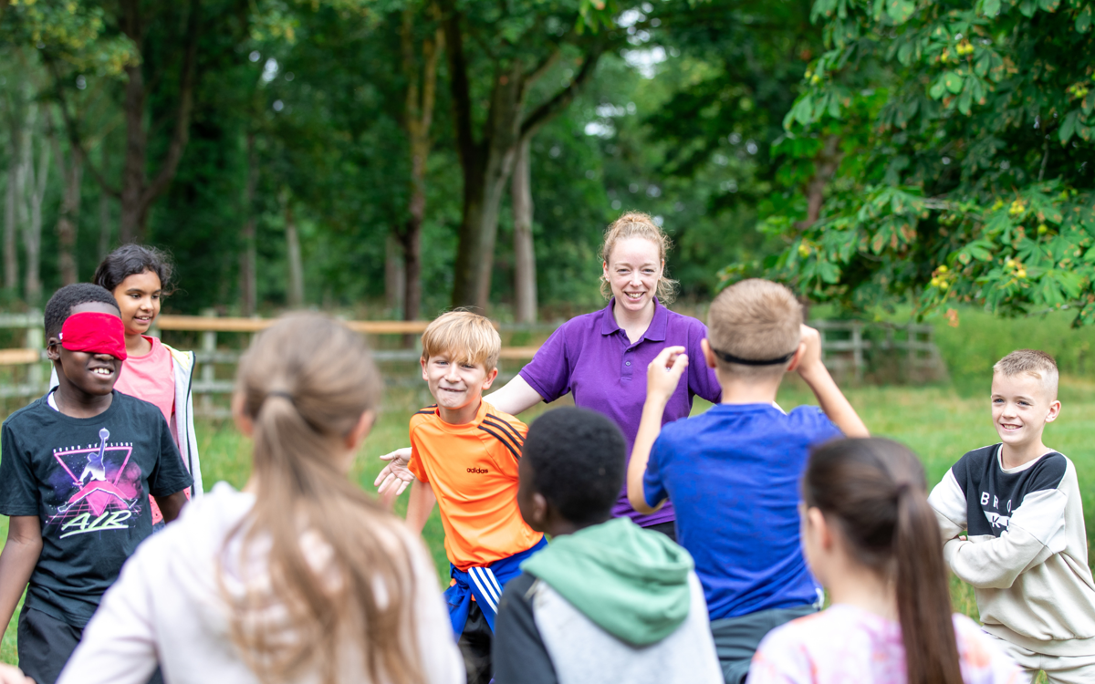 Children and a leader playing a game in a grassy parkland. Two of the children are wearing red blindfolds, running around inside a circle created by their class mates holding their arms out to their sides.