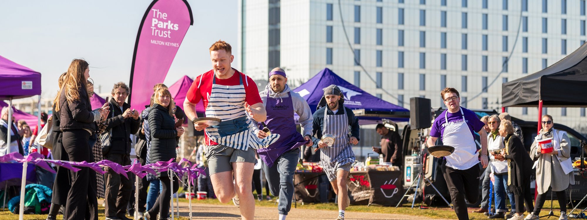 Runners competing in a race holding a frying pan and wearing an apron, with spectators cheering them on