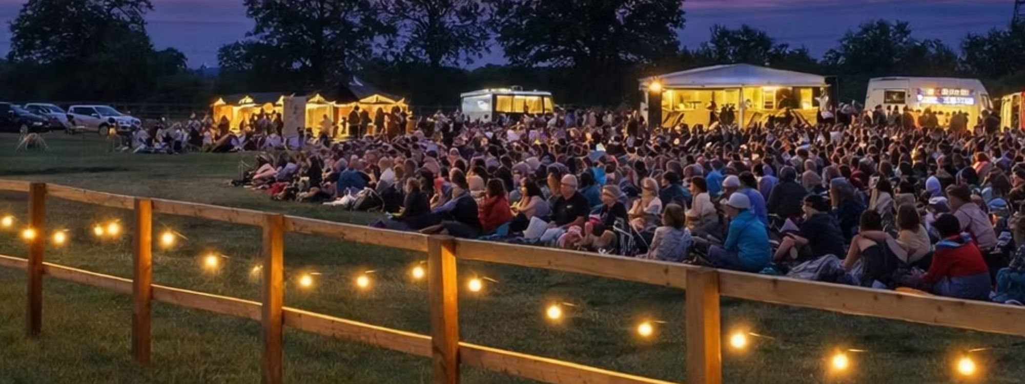 Crowd of people sitting on the grass in a park. it is evening and the site is lit with fairy lights and the lights from the food vendors. 