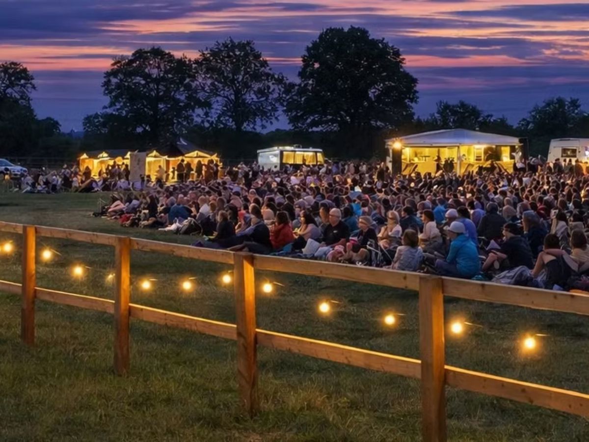 Crowd of people sitting on the grass in a park. it is evening and the site is lit with fairy lights and the lights from the food vendors. 