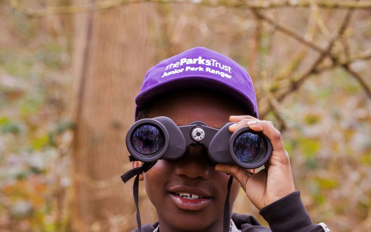 A child in a purple hat is looking into the camera with a pair of black binoculars. He has a smile on his face and there are large textured trees behind him.