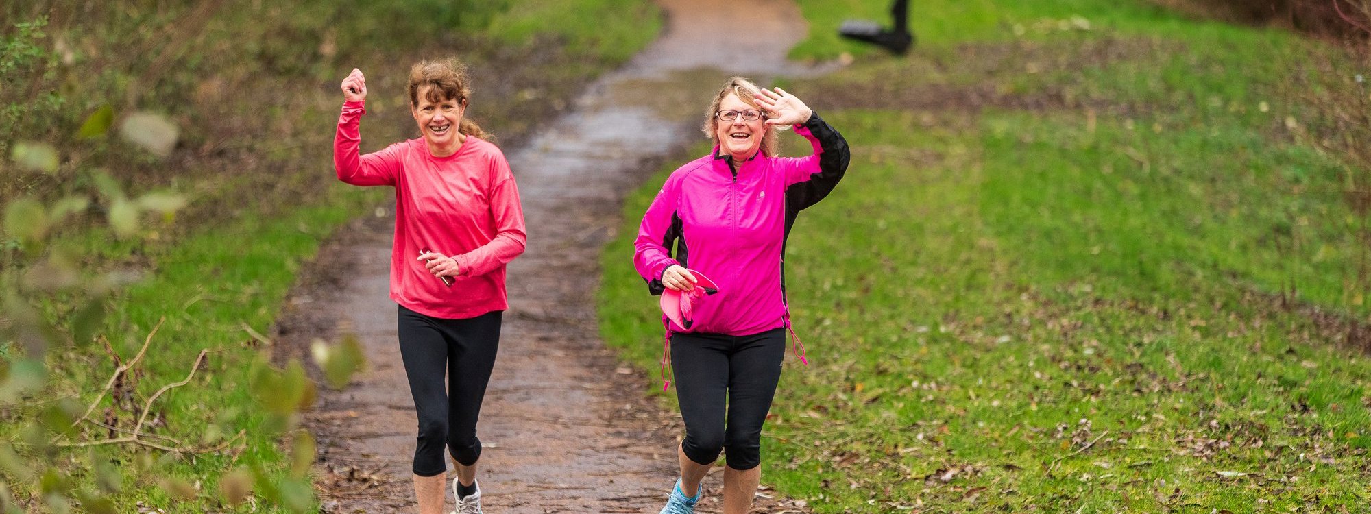 Two female runners waving and smiling in green parkland