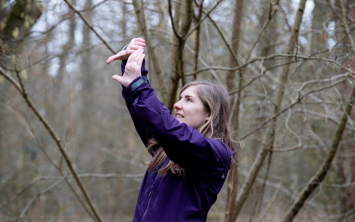 Female in purple jacket holding up hands in a rectangle and looking through in woodland