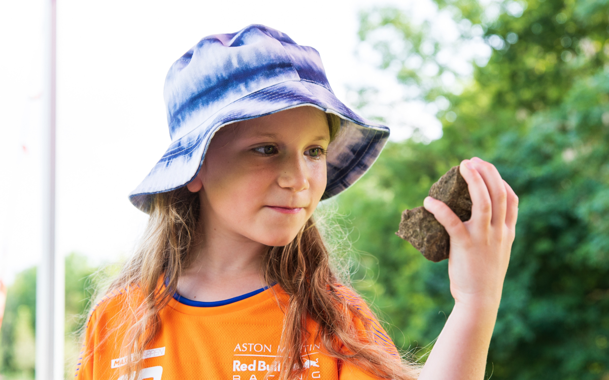 A child in a bright orange t-shirt and blue summer hat is holding a large rock in her hand. She is holding the rock up to her face and smiling.