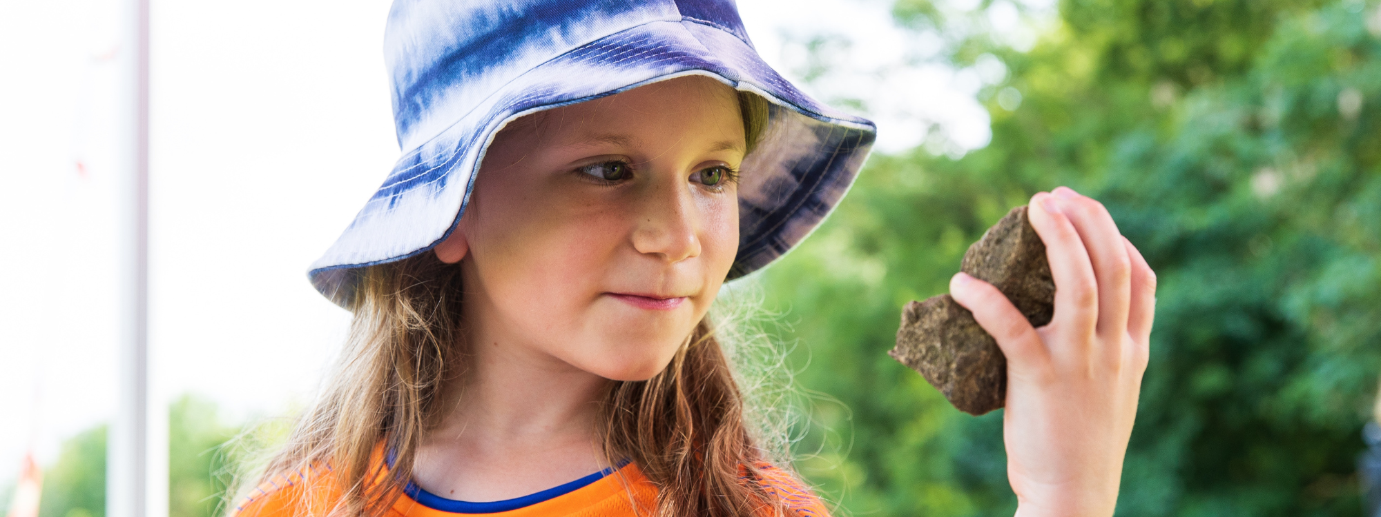 A child in a bright orange t-shirt and blue summer hat is holding a large rock in her hand. She is holding the rock up to her face and smiling.