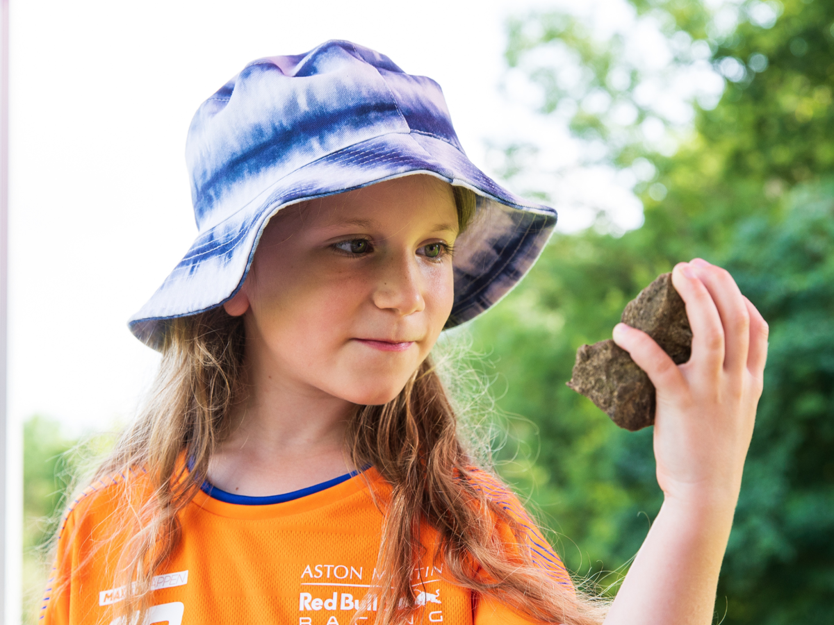 A child in a bright orange t-shirt and blue summer hat is holding a large rock in her hand. She is holding the rock up to her face and smiling.
