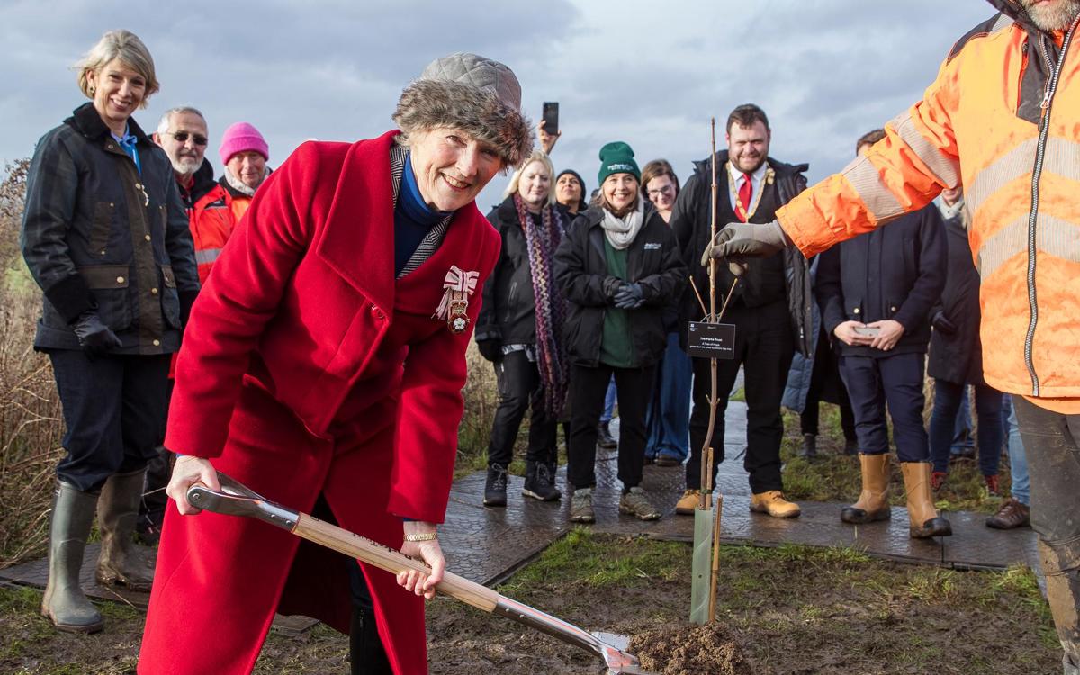 Countess Howe in red coat holding spade with mud in next to tree and group of people behind