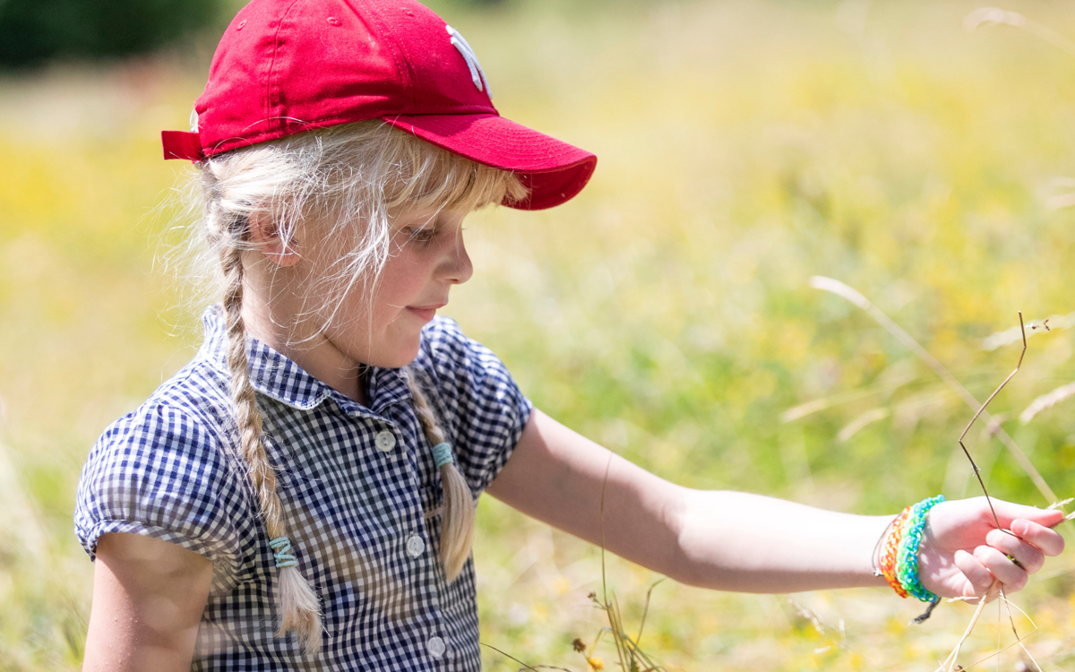 A young girl sitting in a sunny field of long dried grass, holding some in her hand. She has long blonde hair in plaited ponytails and is wearing a red baseball cap and blue chequered school dress.