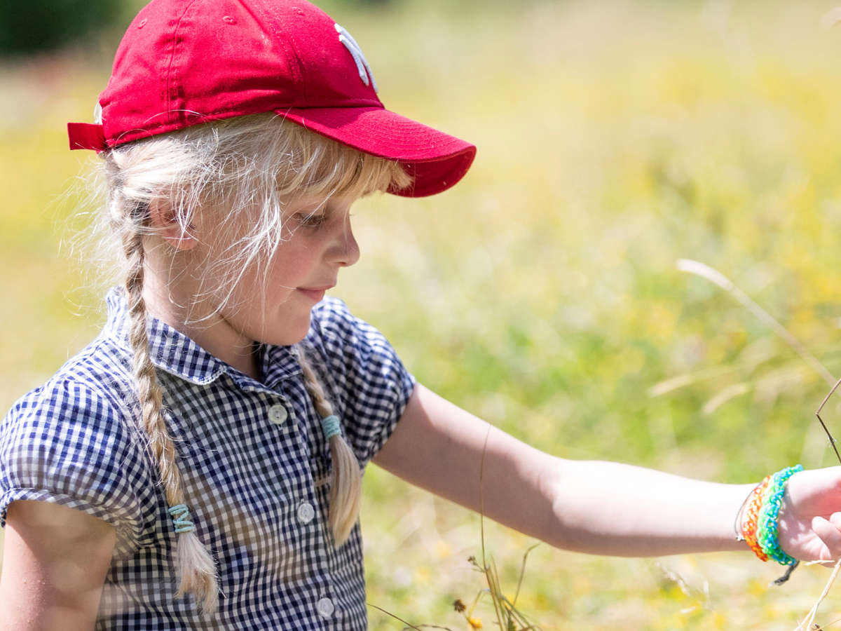 A young girl sitting in a sunny field of long dried grass, holding some in her hand. She has long blonde hair in plaited ponytails and is wearing a red baseball cap and blue chequered school dress.