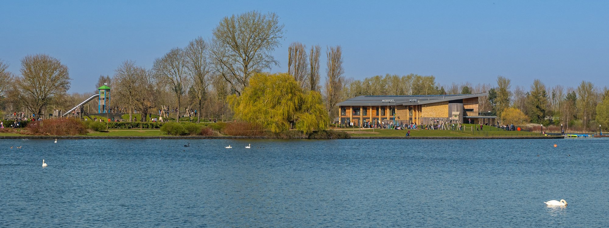 View across Willen Lake looking towards the large water sports centre and outdoor play area on the other side, on a sunny day.
