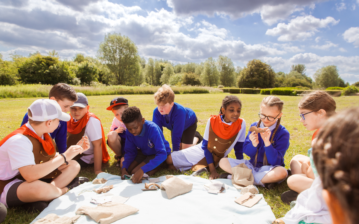 Children sitting in a field in the sunshine studying Roman artefacts. Some are wearing blue school jumpers, others are wearing Roman costumes