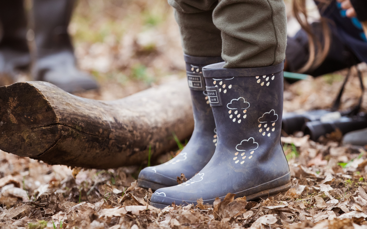 A pair of legs wearing black wellies and grey trousers. The person is stood by a log with lots of brown fallen leaves around.