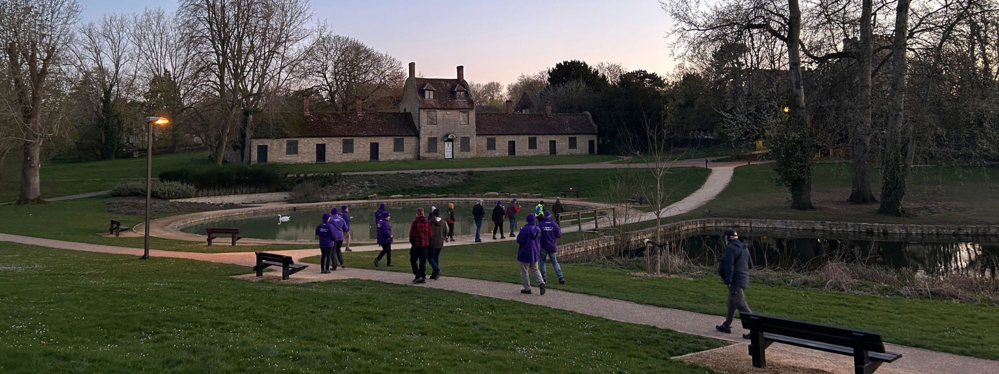 A group walking at dusk through Great Linford Manor Park with formal ponds and buildings in the background. 