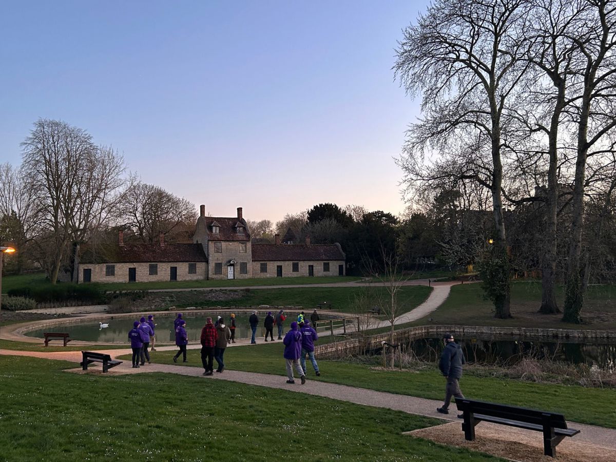 A group walking at dusk through Great Linford Manor Park with formal ponds and buildings in the background. 