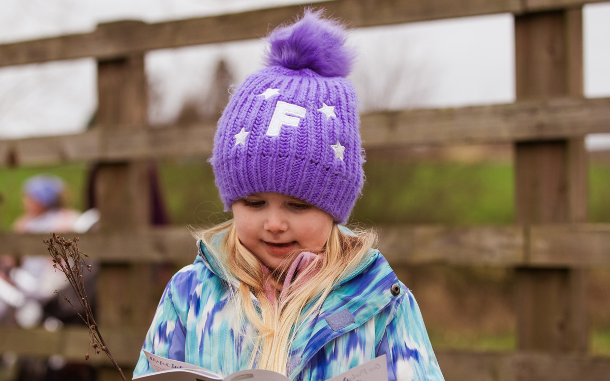 A young girl is stood outside wearing a purple woolly hat and a winter jacket. She is looking at a book and plant that she is holding.