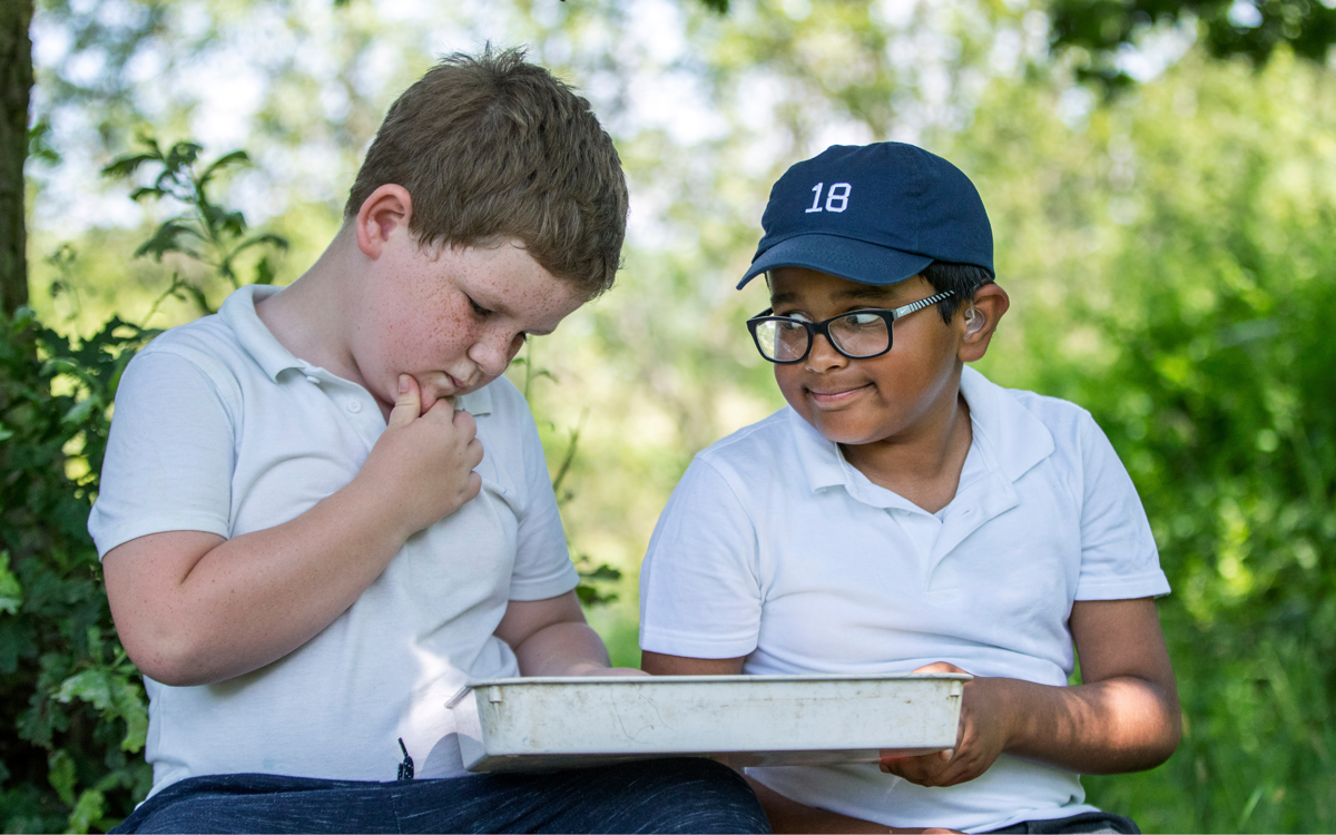 Two boys are sat in the shade under some trees. They are looking at something in a white plastic tray. One boy holds his chin quizzically, the other is looking at the other boy with a cheeky grin.