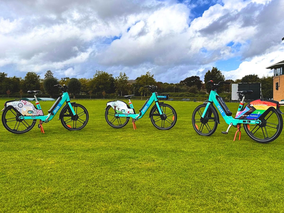 The image shows three bright blue rental bikes with large black wheels, standing in a row on a well-manicured green lawn under a partly cloudy sky. The bikes feature Santander branding, colorful designs on the rear fenders, and are equipped with red kickstands. A clock tower and scoreboard can be seen on the right side of the background, suggesting a sports field or park setting.