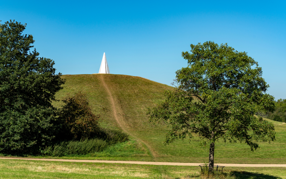 A large white pyramidal art sculpture on top of a big grassy hill. The sky is bright blue and sunny.