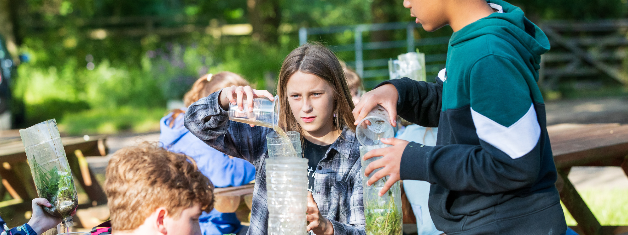 Three teenagers are sat at a bench outside making natural water filters. They are holding plastic cups of dirty water and pouring them through homemade water filters made of plastic bottles that contain leaves, grass and moss. 