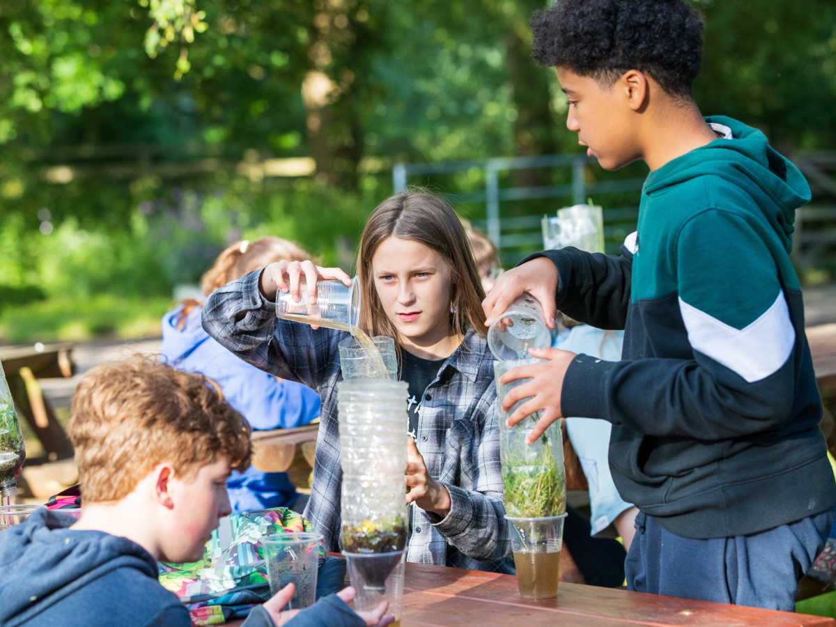 Three teenagers are sat at a bench outside making natural water filters. They are holding plastic cups of dirty water and pouring them through homemade water filters made of plastic bottles that contain leaves, grass and moss. 