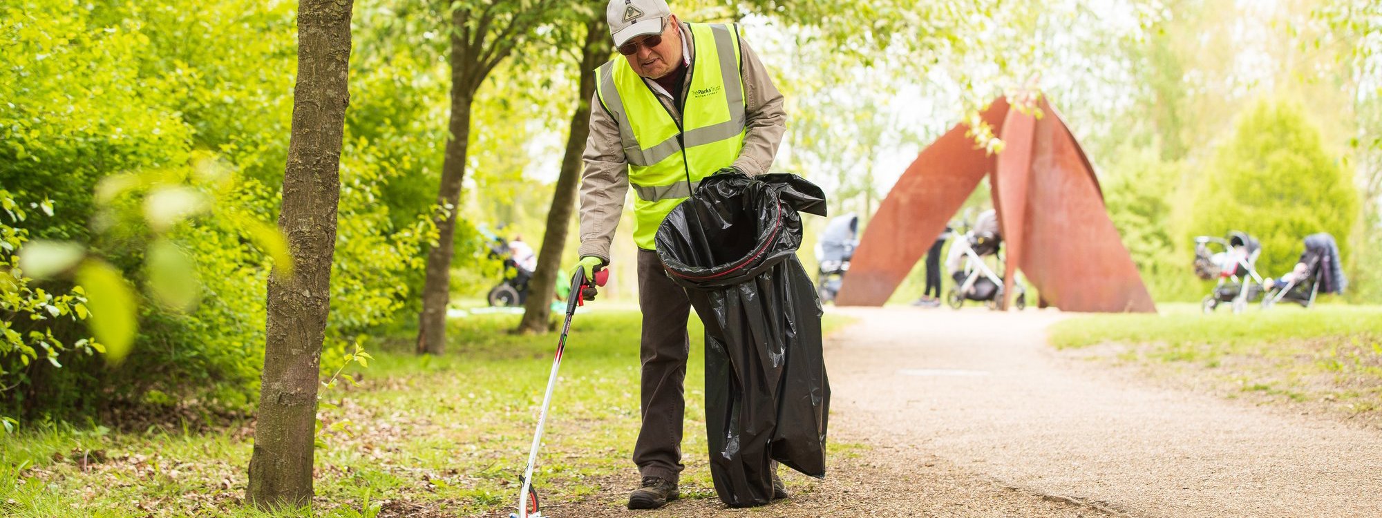 A volunteer in hivis litter picking in a parkland scene with a large public artwork in the background