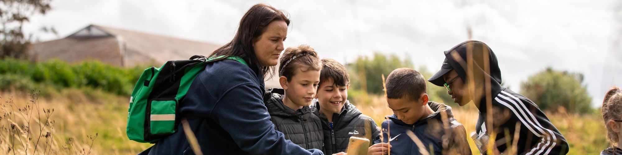 Teacher leading session with children in park. The female teacher is taking a photo on her phone, while standing in long brown grass. Four young boys are looking at the teacher's phone and smiling.
