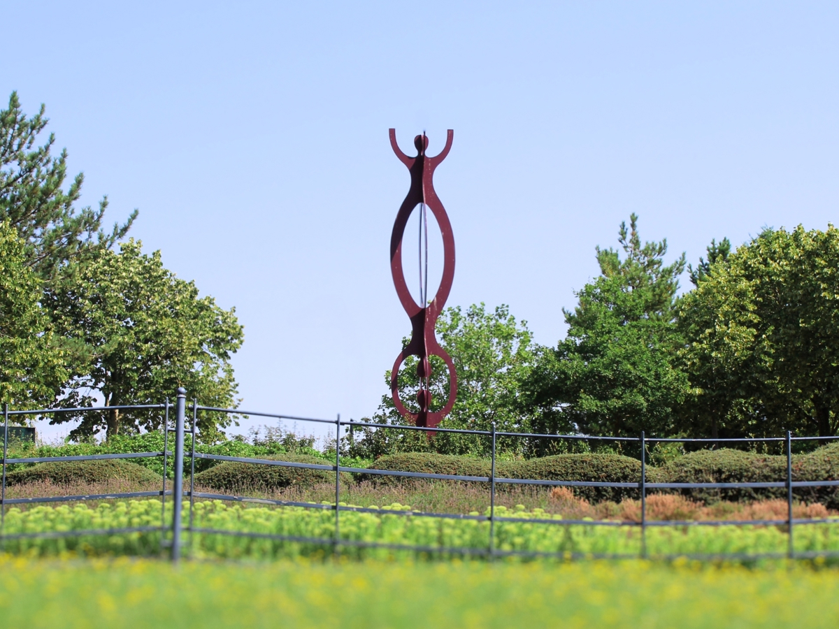 The image shows a tall, abstract metal sculpture with a deep reddish hue, prominently displayed in a landscaped area. The sculpture features two elongated, interconnected loops designed to look like people. The area surrounding the sculpture is fenced off with a black metal railing, and the ground is covered with low-lying greenery and small flowers. In the background, there are tall, leafy trees under a clear blue sky, creating a peaceful and well-maintained park-like setting.